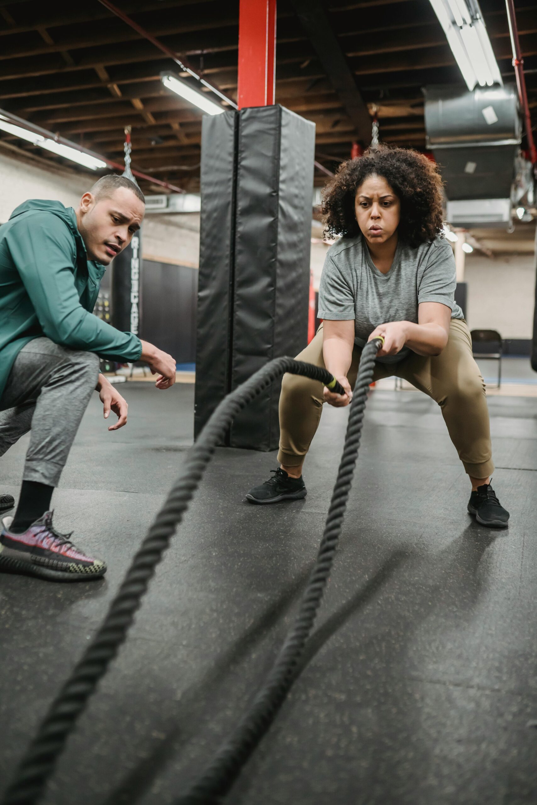 Strong African American female in activewear training with battle ropes near fit male coach in fitness center